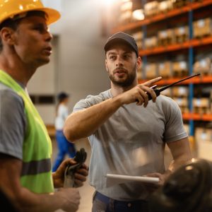 Young dispatcher communicating about delivery schedule with warehouse worker in factory warehouse.